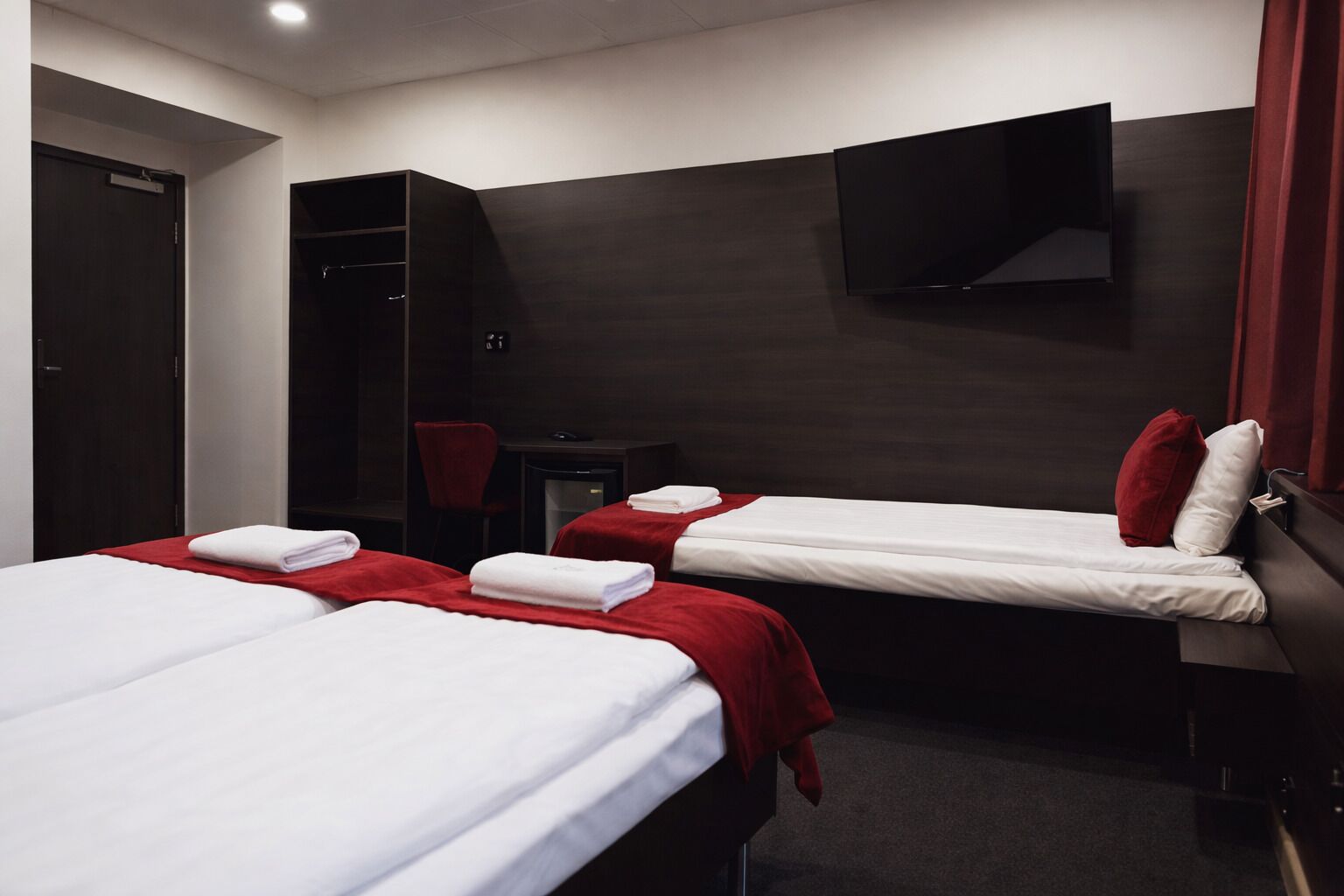 A hotel room with two beds, each with white linens and red bedspreads. There is a desk with a chair, a television mounted on the wall, and a wardrobe.