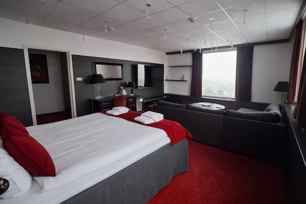 A modern hotel room with a large bed, a sofa, a desk, and a large window.