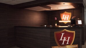 Een receptiebalie met donkerhouten afwerking, een logo met de tekst 'LH' en 'Liljeholms Stadshotell', een kleine potplant en een digitaal scherm.