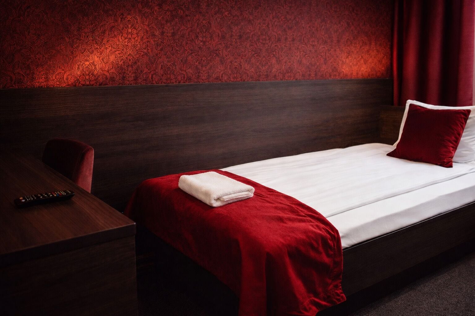 Habitación de hotel con una cama individual, una mesita de noche de madera, una manta roja y un control remoto sobre la mesita.