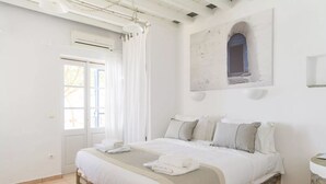 Une chambre avec un lit dont la base est en pierre, du linge de lit blanc, une grande fenêtre avec des rideaux et une œuvre d’art accrochée au mur.