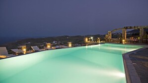 Une piscine entourée de chaises longues et illuminée, bordée de constructions en pierre et offrant une vue sur le paysage.