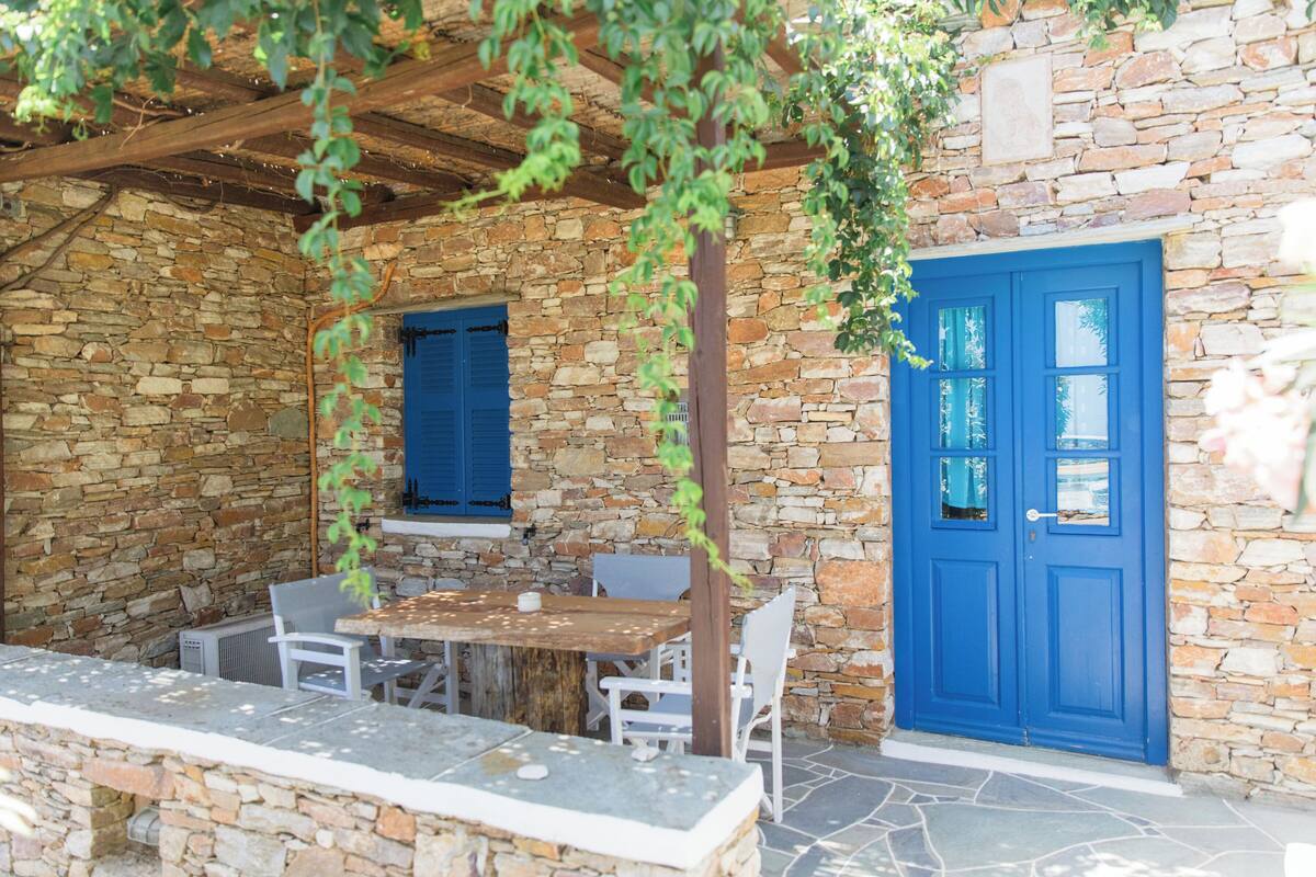 Ein Steinhaus mit einer Holzpergola, einer blauen Tür und einem Tisch mit Stühlen.