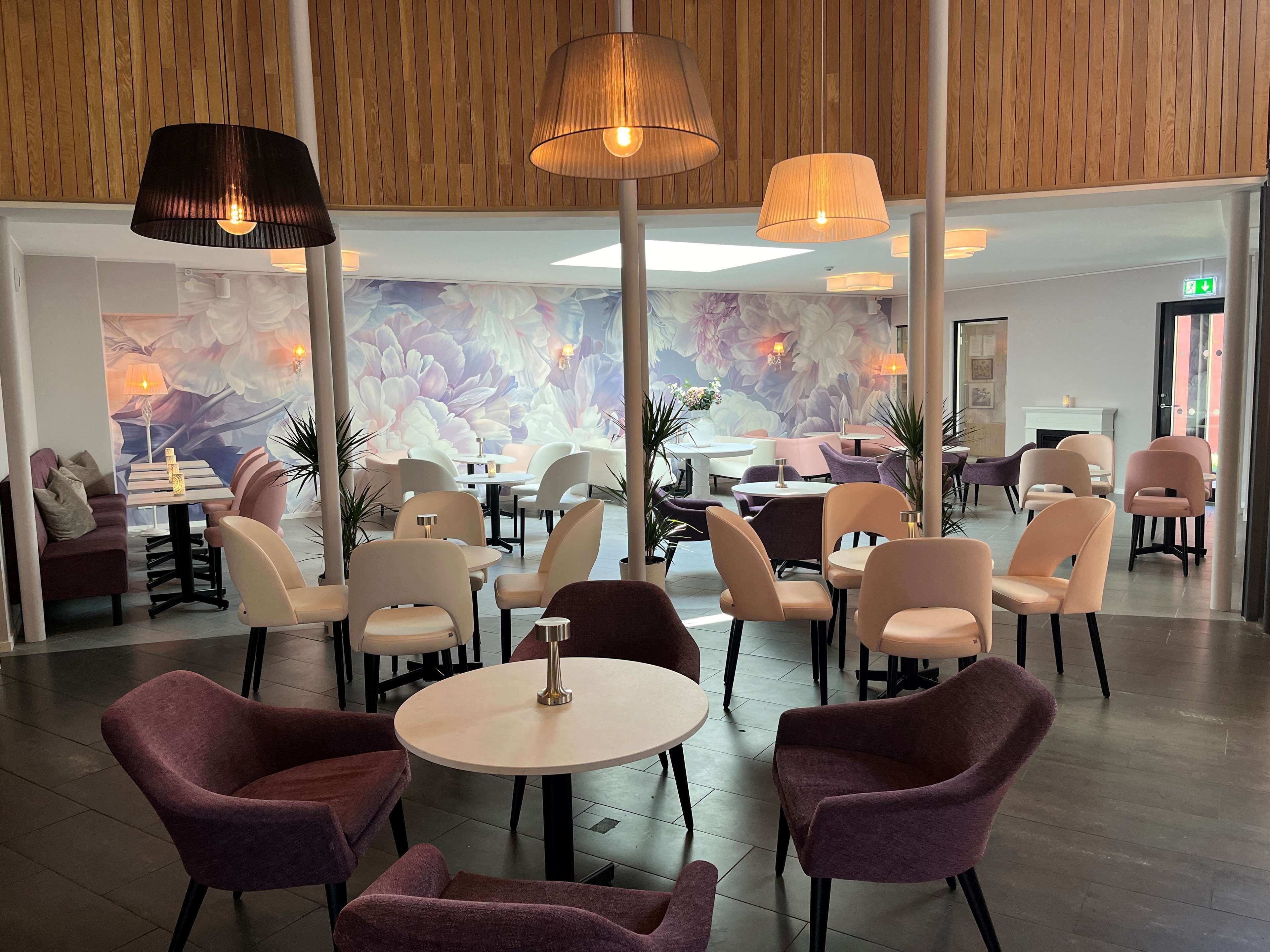 A modern hotel lobby with round tables and chairs, wooden paneling on the ceiling, and a floral mural on the wall.