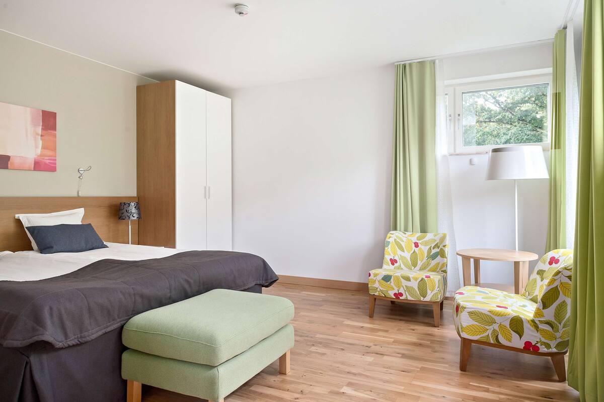 Un dormitorio con cama, un banco verde, dos sillones estampados, una mesita redonda y una ventana con cortinas.