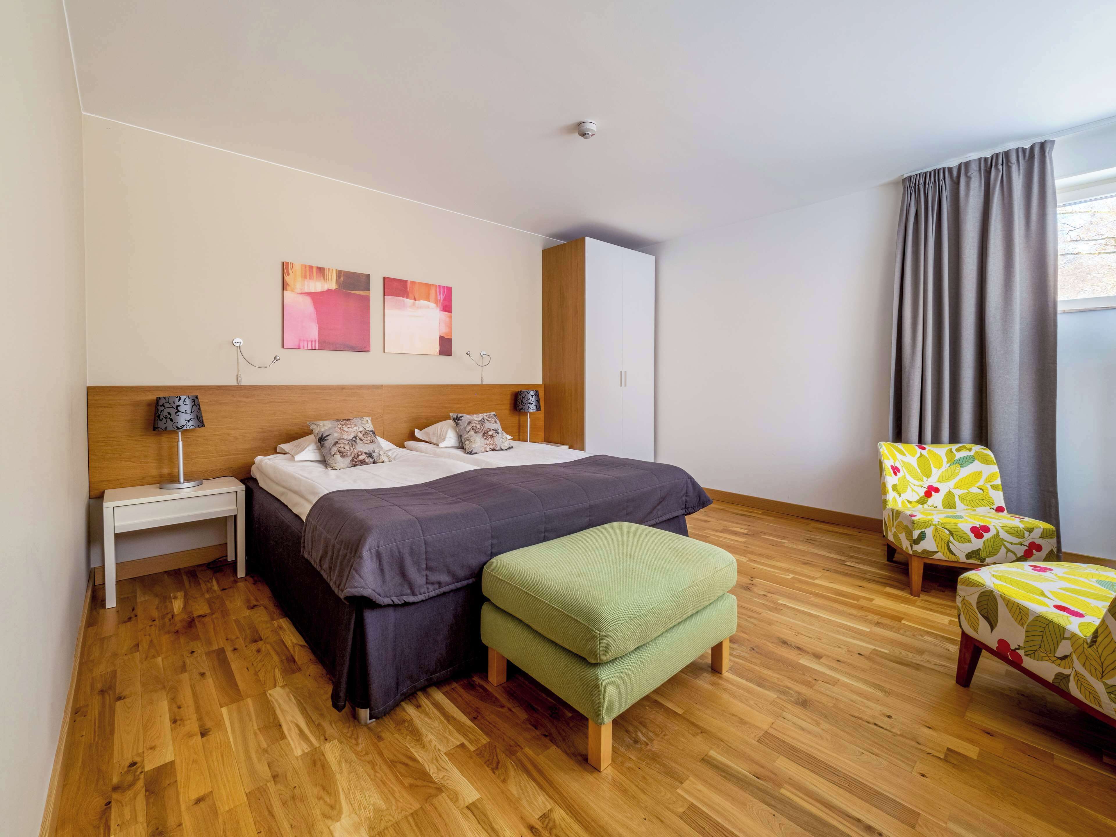 Une chambre d’hôtel moderne avec un plancher en bois, un lit recouvert d’une couette grise, un pouf vert, un fauteuil à motifs floraux et une fenêtre avec des rideaux gris.