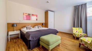 A modern hotel room with a wooden floor, a bed with a grey duvet, a green ottoman, a floral armchair, and a window with grey curtains.