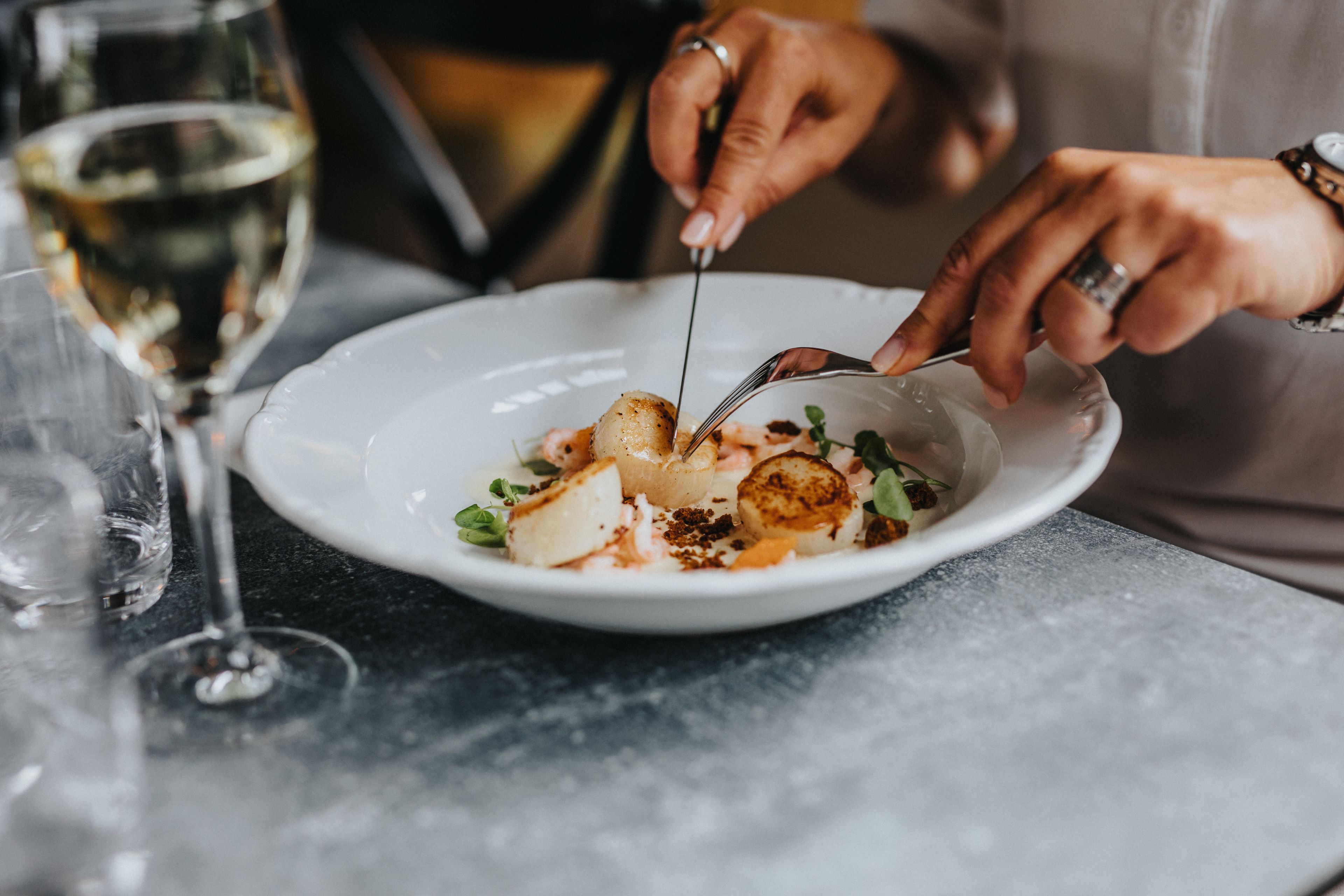 A person dining at a table with a white plate of food, a glass of white wine, and a fork.