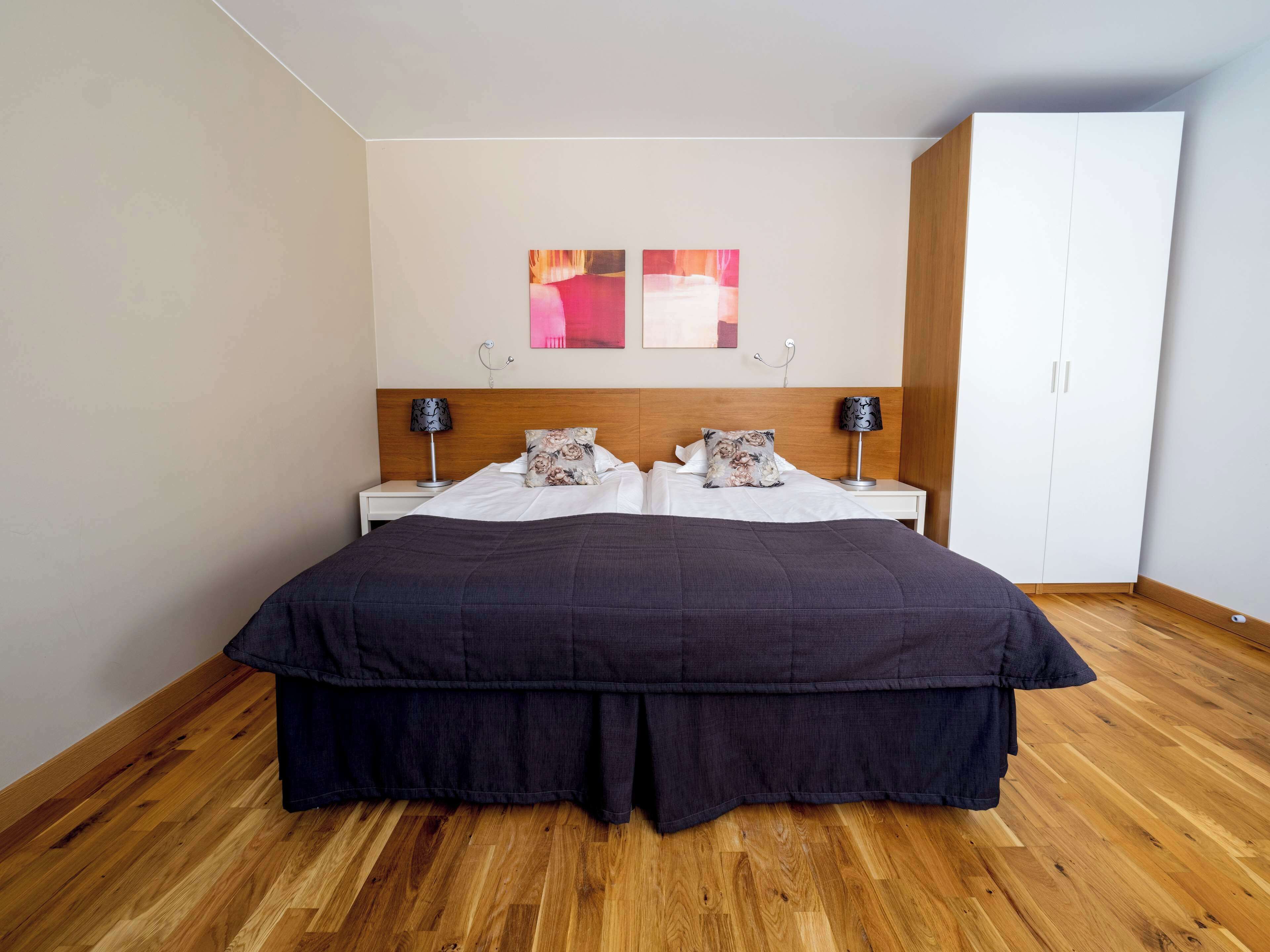 Un lit double avec une couvre-lit sombre, deux tables de chevet avec des lampes, une tête de lit en bois, une armoire blanche et un mur orné d’œuvres d’art abstraites.