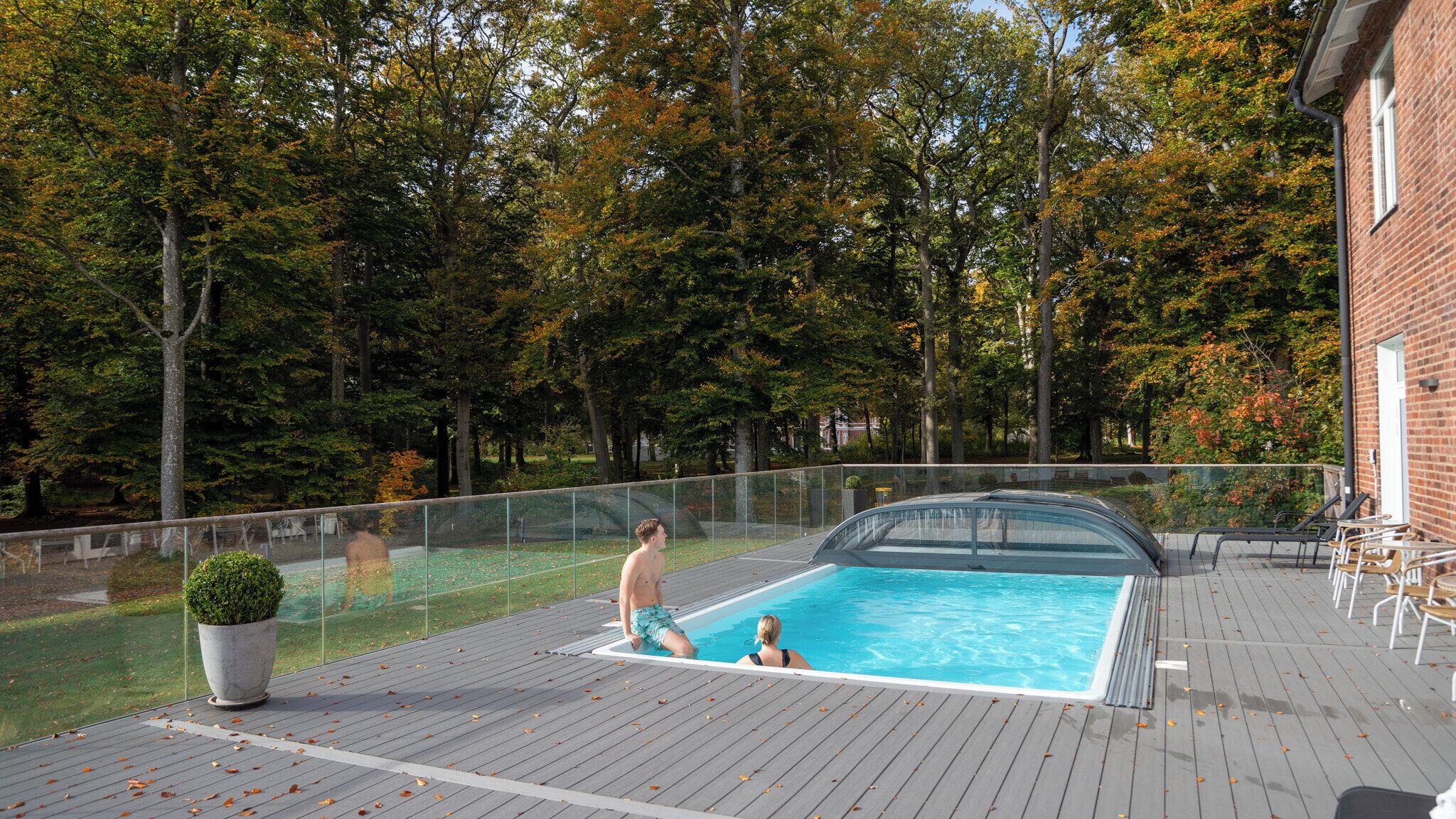 Una piscina con cerramiento de vidrio, rodeada de árboles y una terraza de madera.