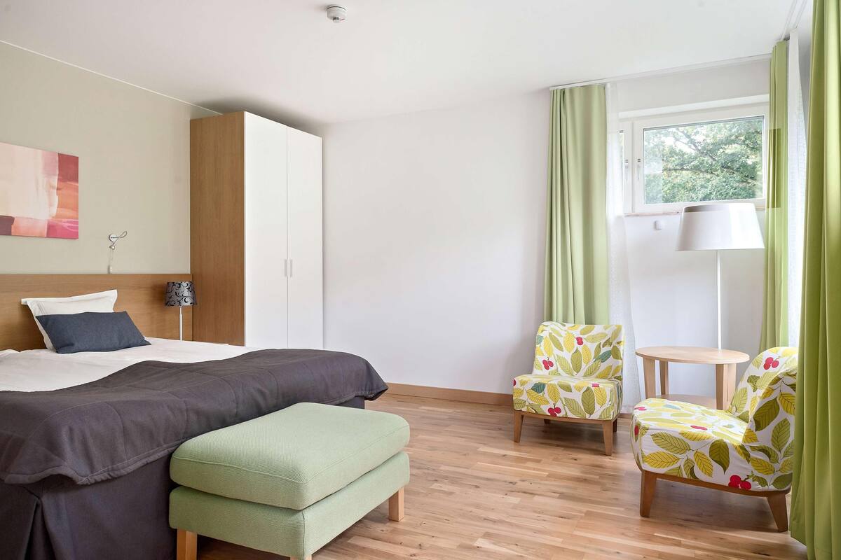Un dormitorio con una cama, un banco, dos sillas, una mesa y un ventanal con cortinas.
