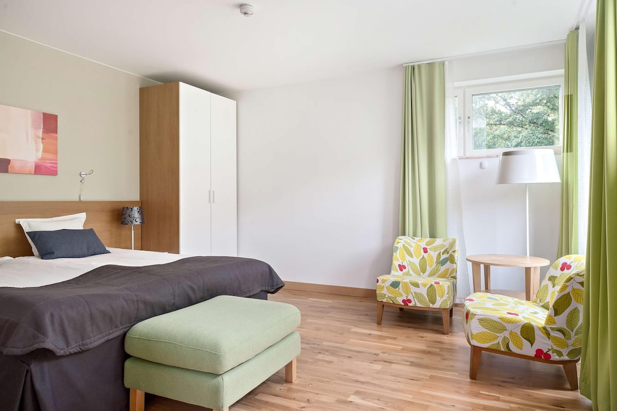 Un dormitorio con una cama, un banco, dos sillas, una mesa y un ventanal con cortinas.