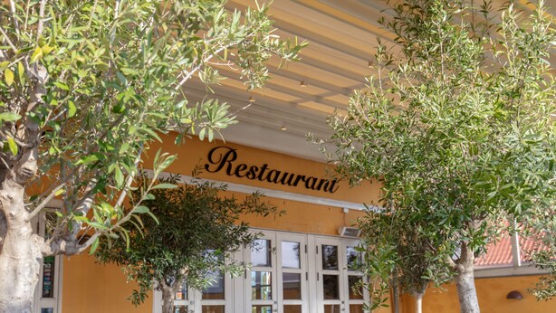 Un área de estar al aire libre con mesas y sillas, un letrero que dice "Restaurant" y árboles que dan sombra.