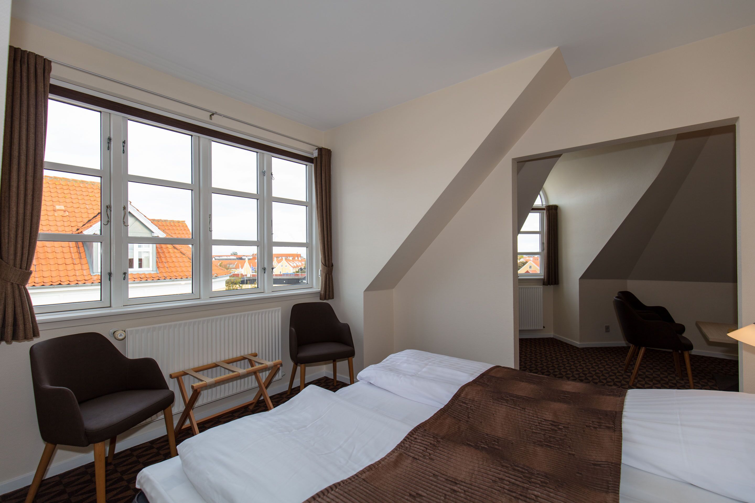 Una habitación de hotel con un ventanal grande, dos sillas, una cama y una mesita.