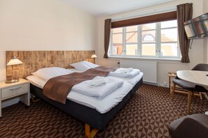 Ein Hotelzimmer mit einem großen Bett, zwei Nachttischen mit Lampen, einem kleinen Tisch mit Stühlen und einem an der Wand montierten Fernseher.