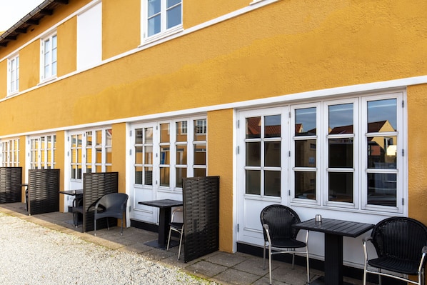 An outdoor seating area with tables and chairs, a gravel path, and a yellow building facade with white-framed windows.