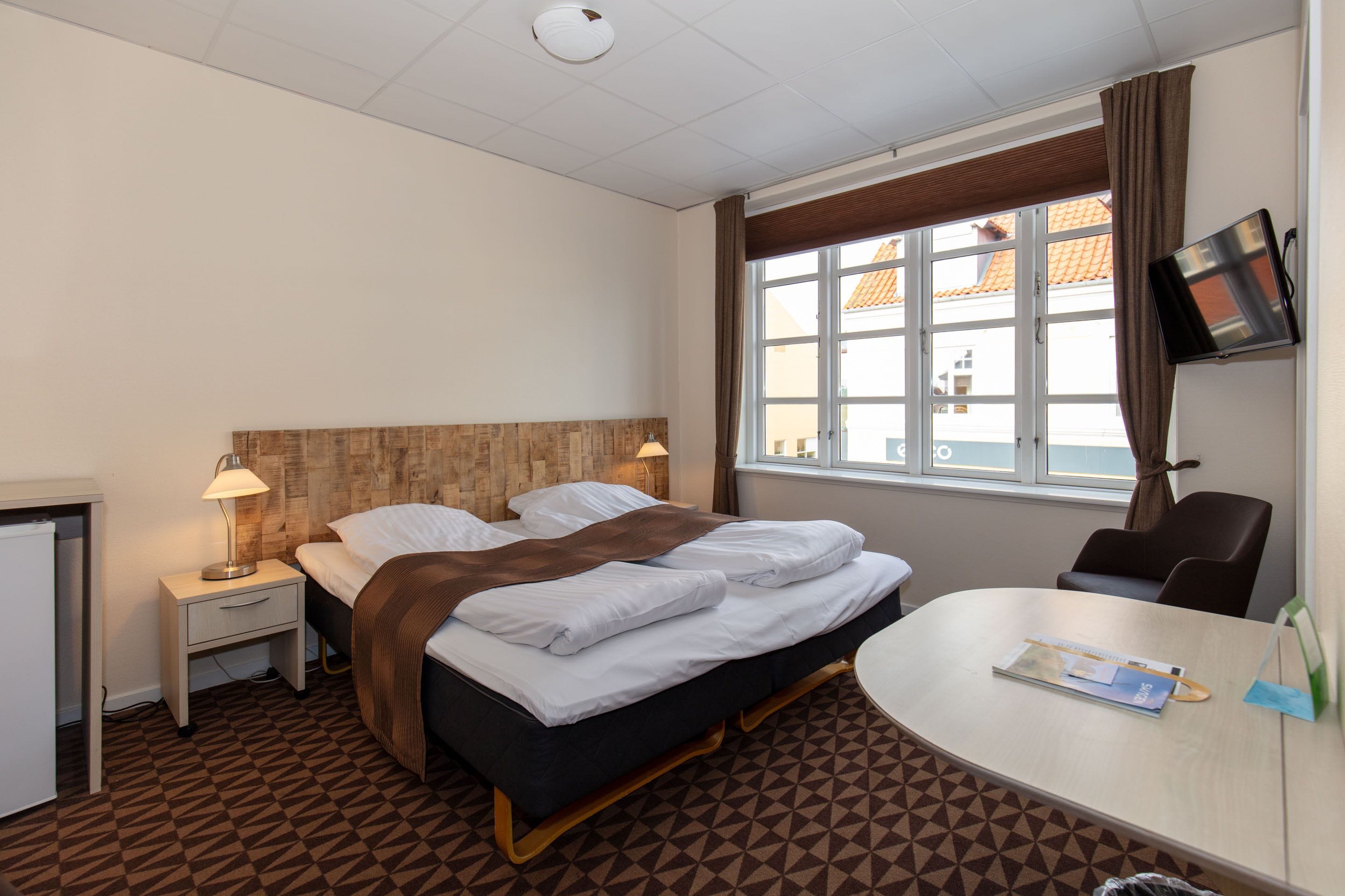 Habitación de hotel con cama, mesita de noche, lámpara, televisor y vistas al exterior.