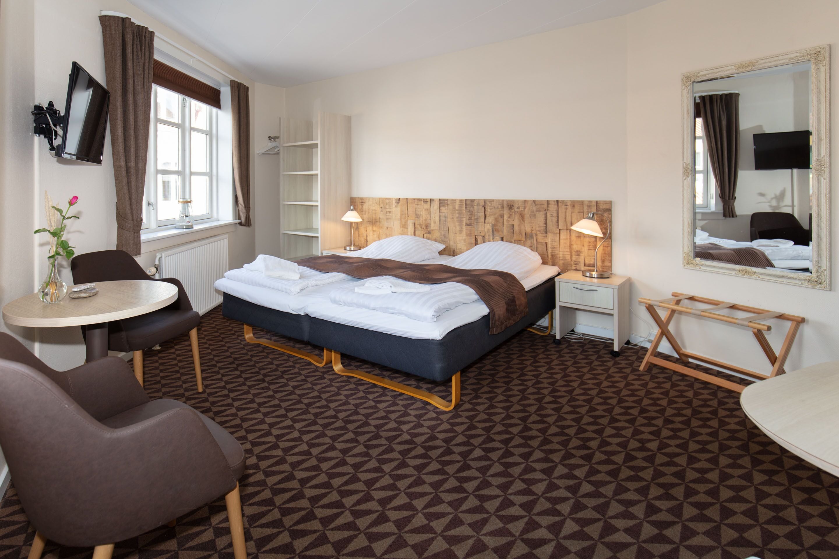 Una habitación de hotel con cama, mesitas de noche, un escritorio, una silla, un espejo y una ventana con cortinas.