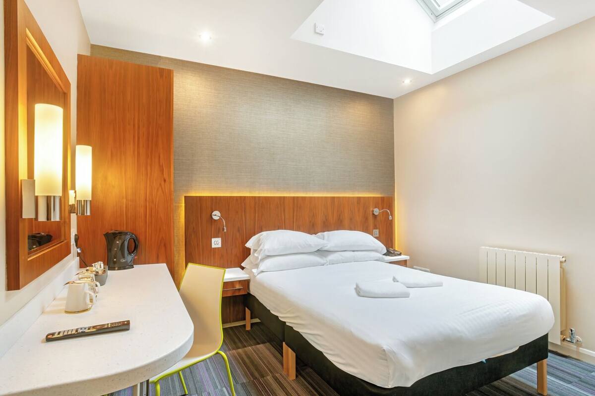 A hotel room with a bed, bedside table, chair, and a skylight.