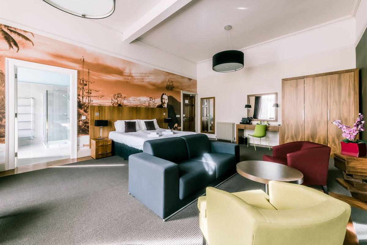 A modern hotel room with a bed, a sofa, a chair, a small table, and a wooden wardrobe.