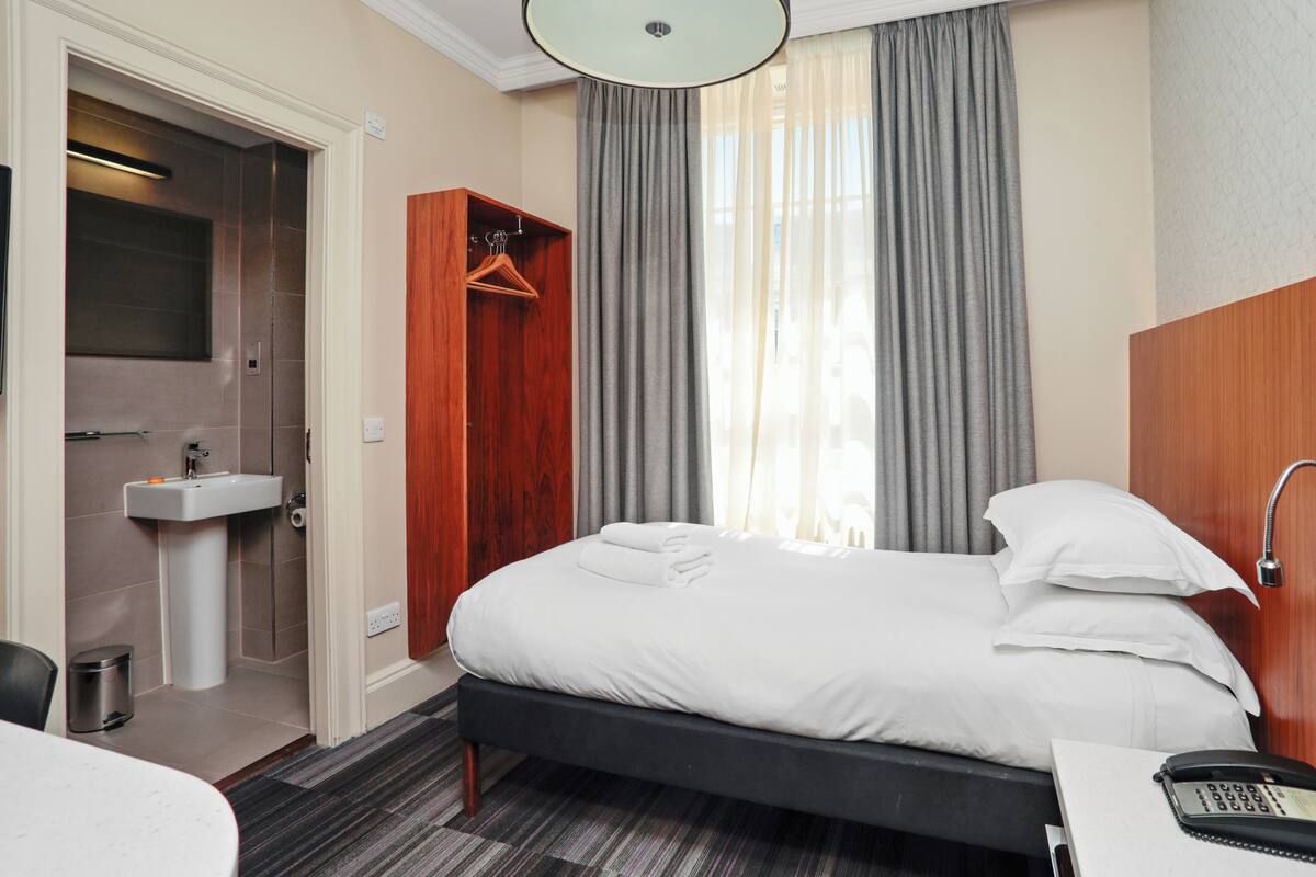 A hotel room with a bed, a bedside table with a phone, a wooden wardrobe, a bathroom with a sink, and a window with curtains.