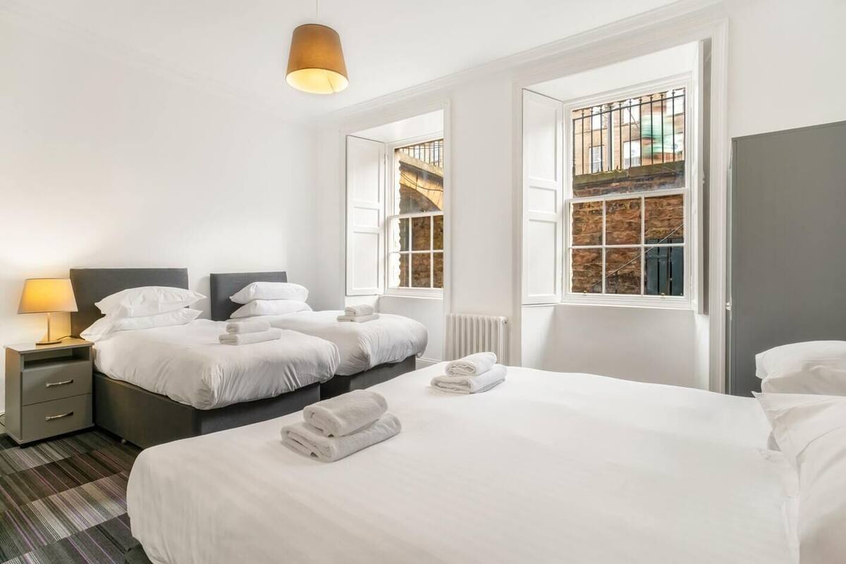 A hotel room with two beds, a nightstand, a lamp, and large windows with a view of a brick building.