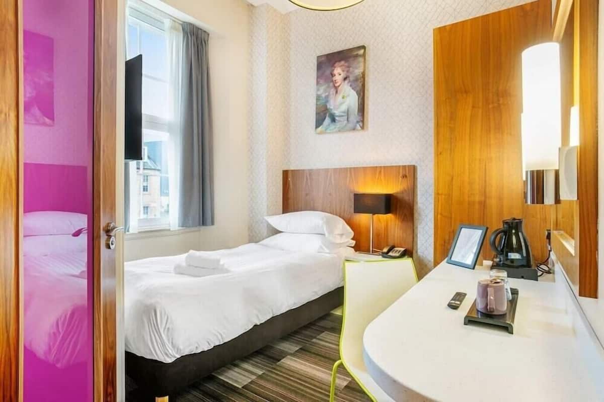 A hotel room with a bed, bedside table, lamp, and a painting on the wall.