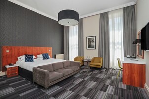 A hotel room with a large bed, a sofa, a desk, and a chair. There is a window with curtains, a painting on the wall, and a lamp on the desk.