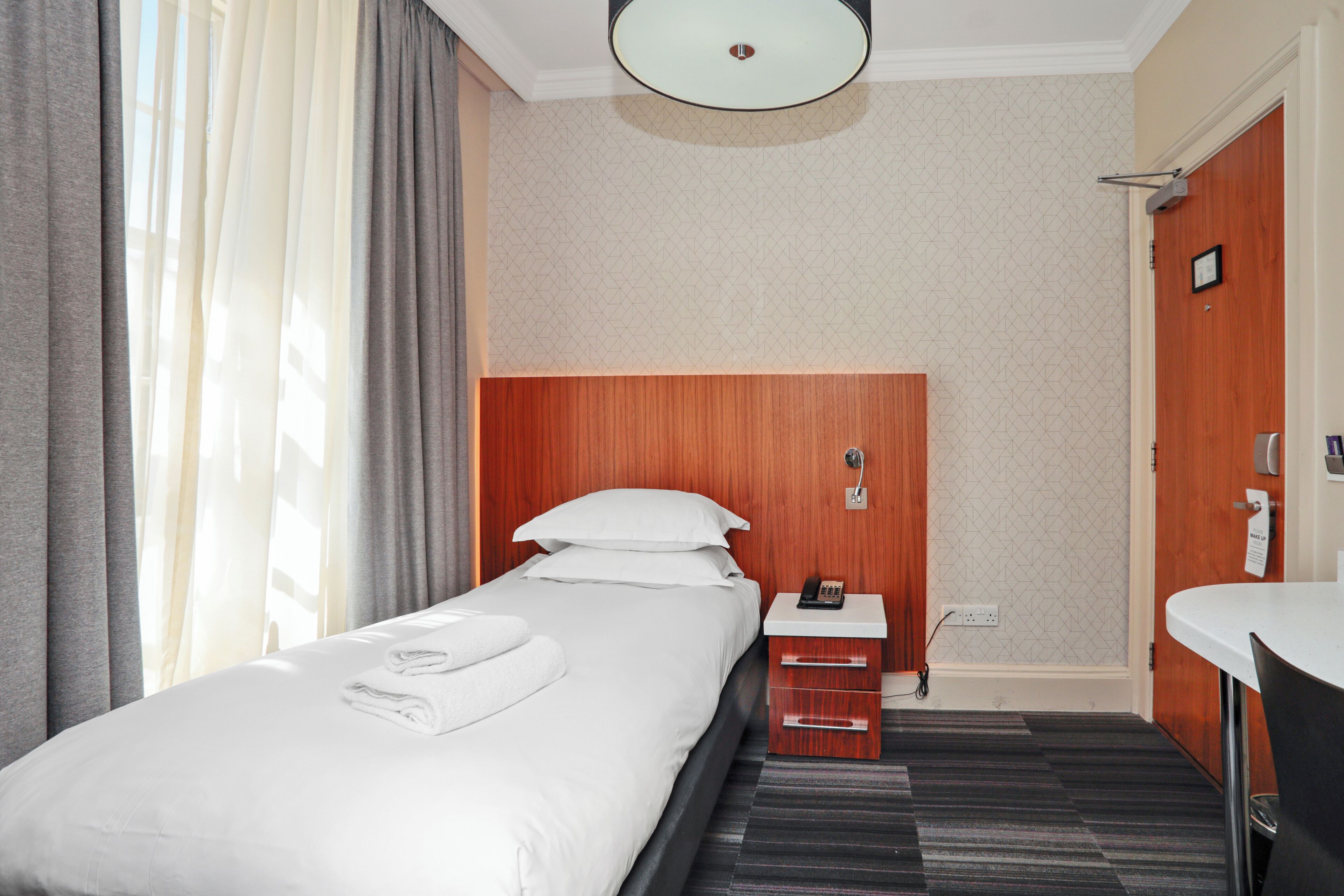 A hotel room with a neatly made bed, a bedside table with a phone, a small drawer unit, a wooden wardrobe, and a bathroom with a sink and mirror.