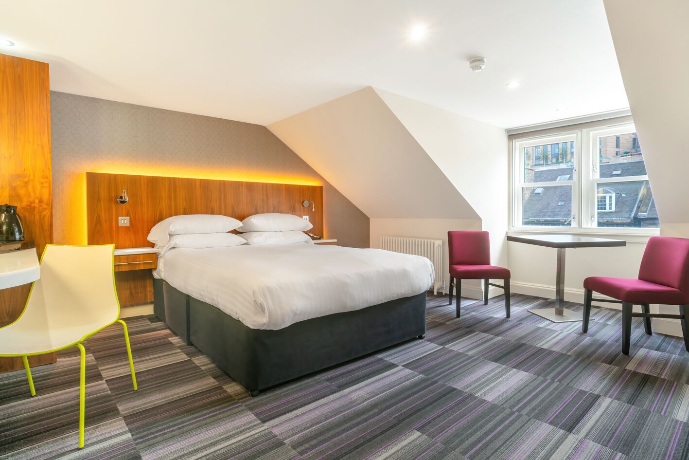 A modern hotel room with a large bed, two chairs, a small table, and a view of the city through a large window.