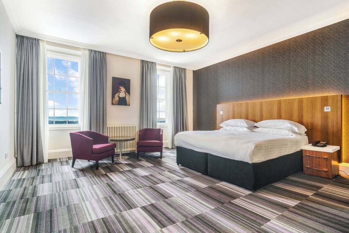 A hotel room with a large bed, two armchairs, a round table, and a view of the outdoors through large windows.