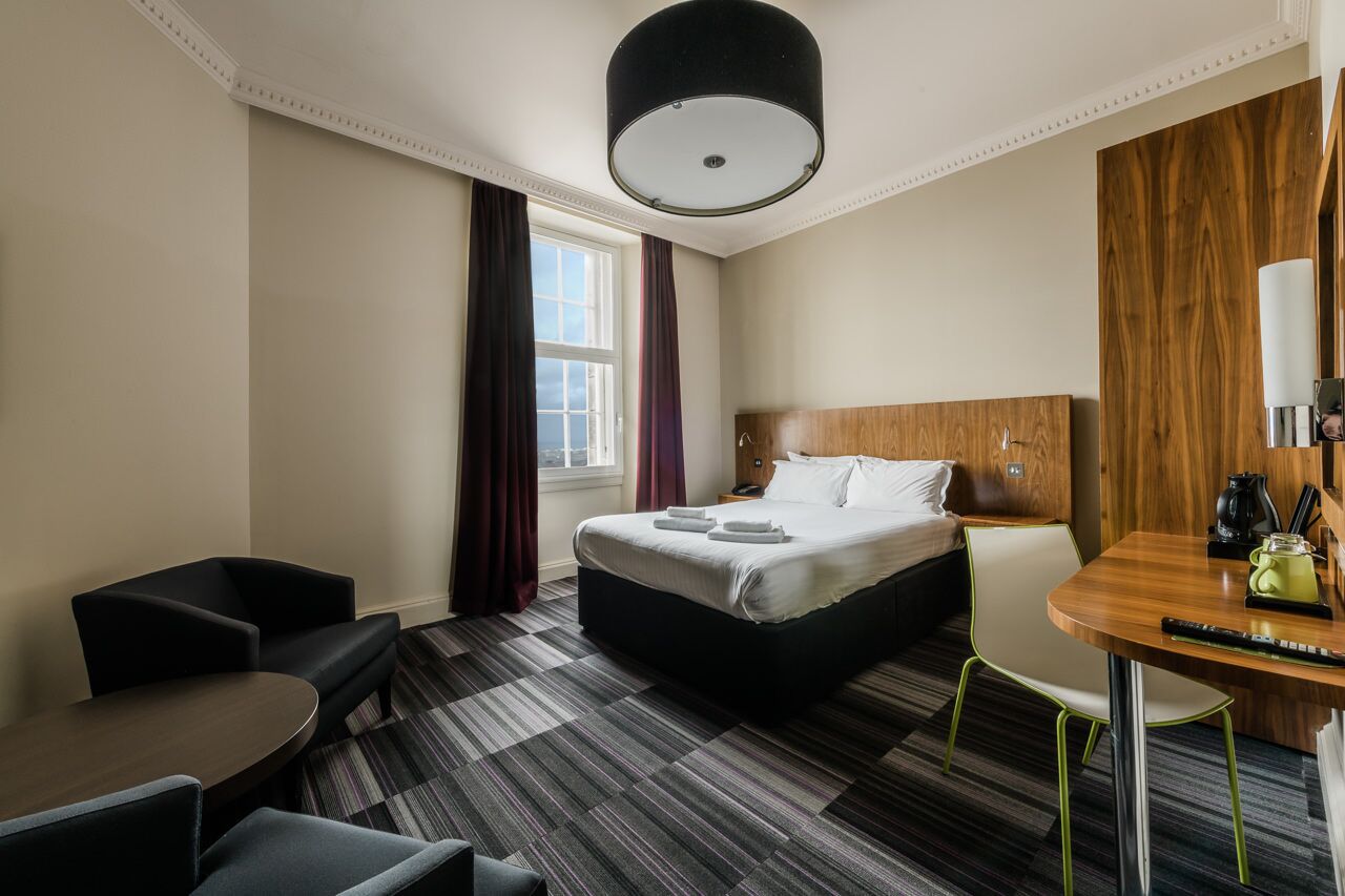 Una camera d'albergo con un letto, una scrivania, una sedia, un tavolino e una finestra con tende.
