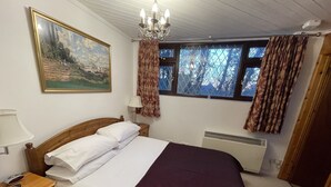 A bedroom with a wooden bed, white bedding, a painting on the wall, a chandelier, a window with curtains, and a bedside lamp. - Tovey Lodge (Hassocks)