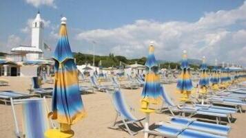 Une plage bordée de rangées de chaises longues et de parasols, un bâtiment blanc surmonté d’une tour, et un ciel dégagé.