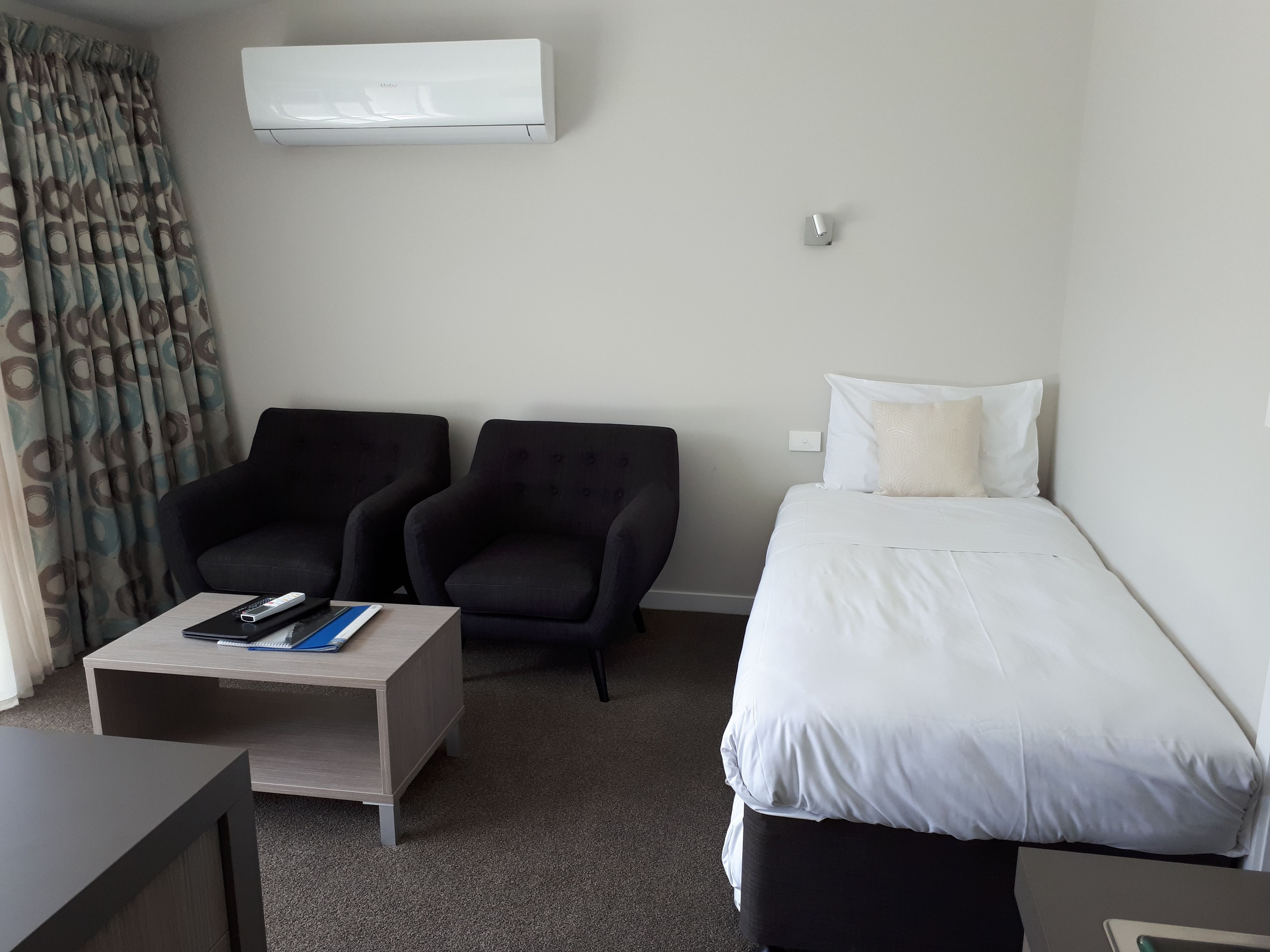 Habitación de hotel con una cama, dos sillones, una mesita con un control remoto y una libreta, y un equipo de aire acondicionado en la pared.
