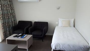 Habitación de hotel con una cama, dos sillones, una mesita con un control remoto y una libreta, y un equipo de aire acondicionado en la pared.