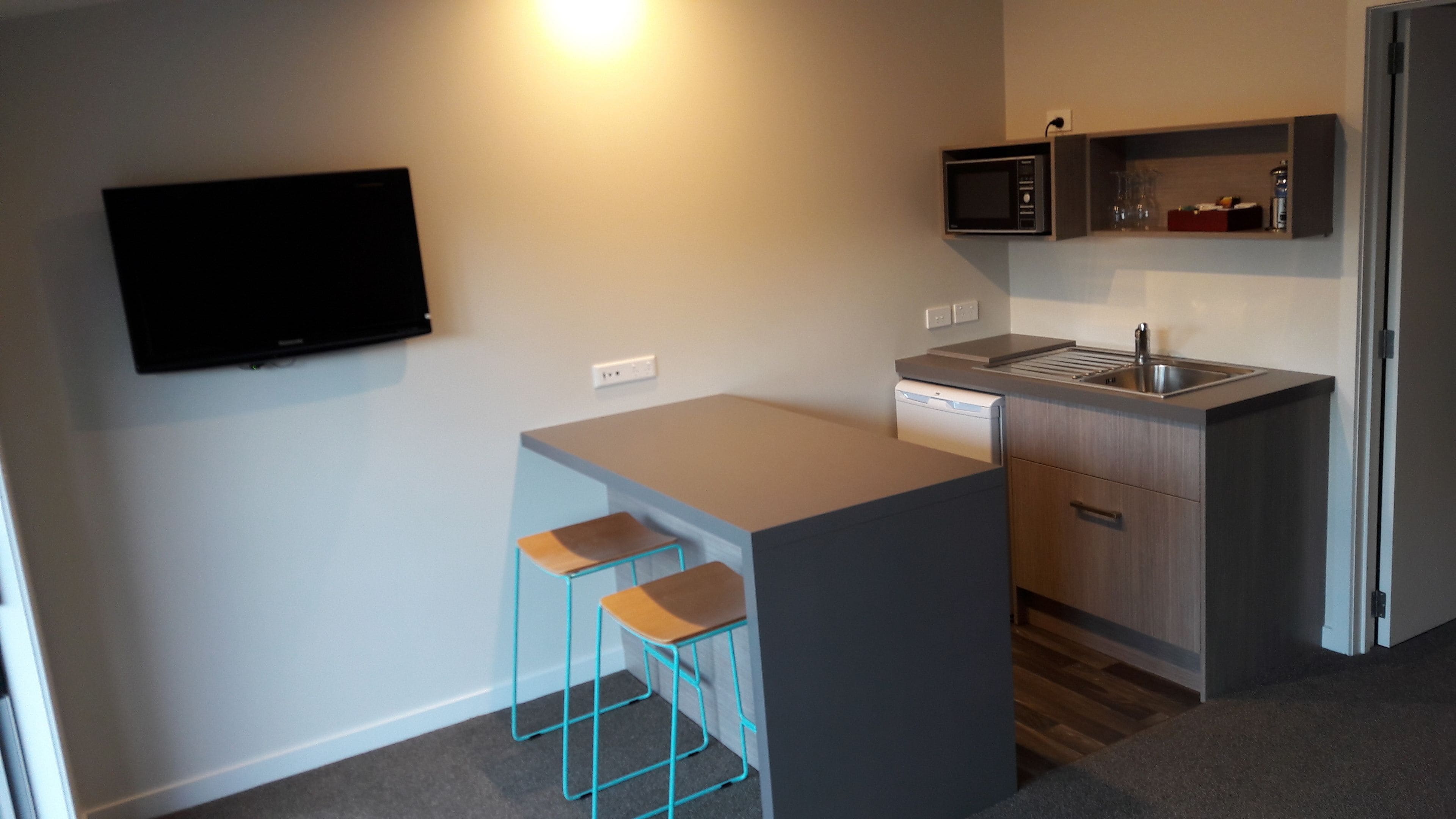 A compact kitchen with a built-in microwave, a small sink, and a dishwasher. There is a flat-screen TV mounted on the wall and a small bar area with stools.