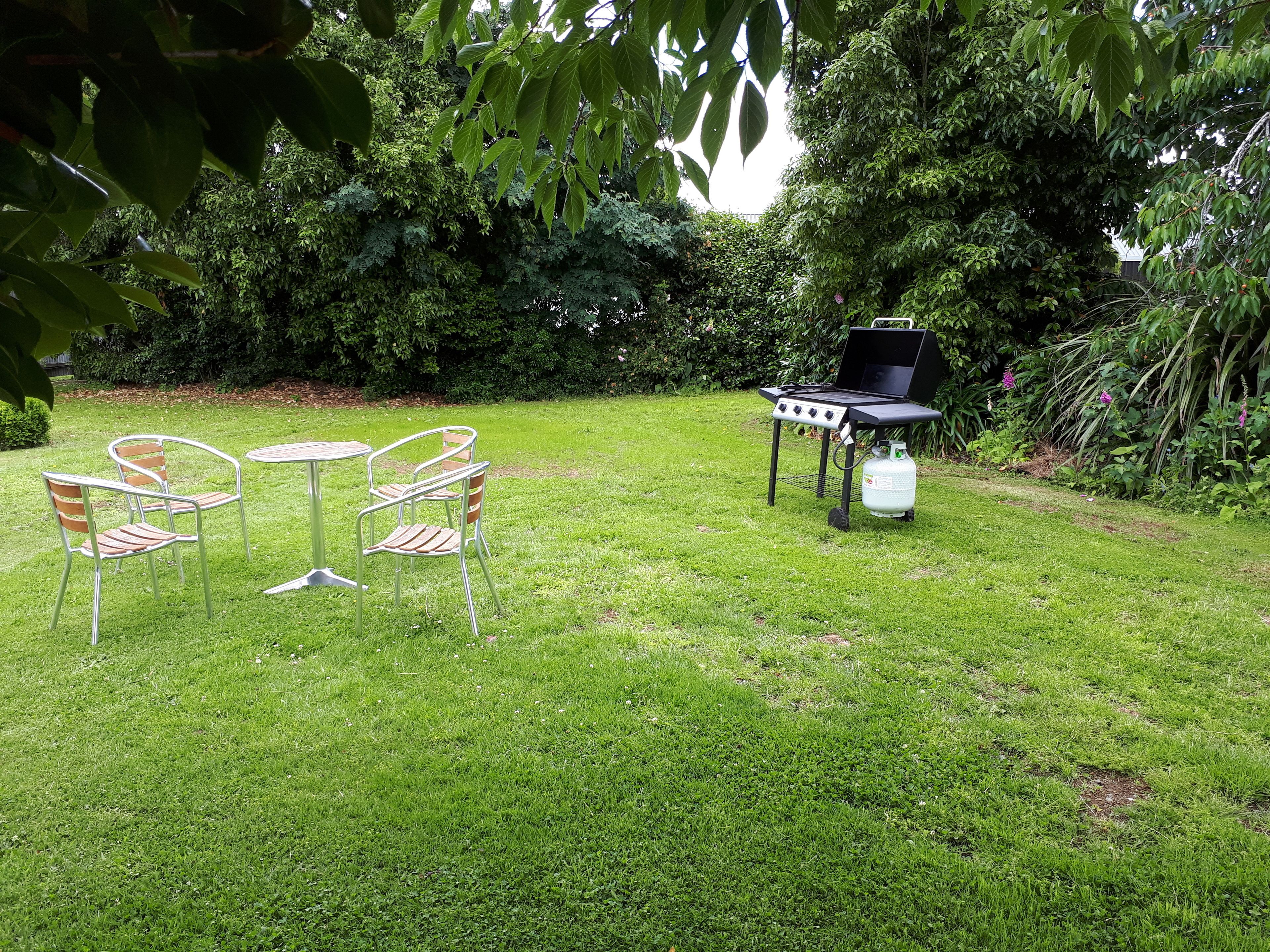 A grassy outdoor area with a barbecue grill, two metal chairs, and a table.