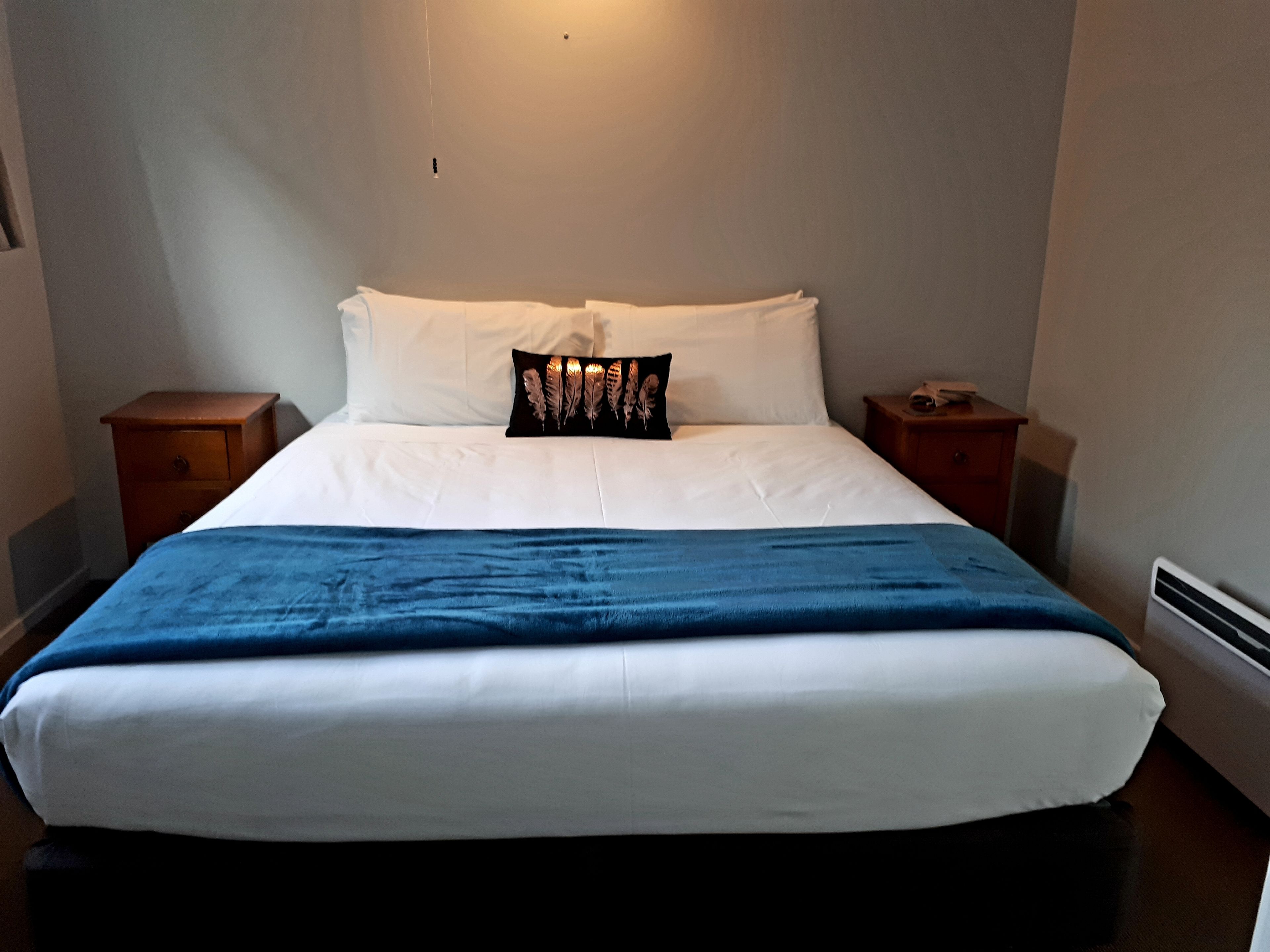 Una cama bien hecha con sábanas blancas y una manta azul. Hay un almohadón negro con un diseño de plumas sobre la cama. Dos mesitas de noche de madera flanquean la cama, cada una con una pequeña lámpara.