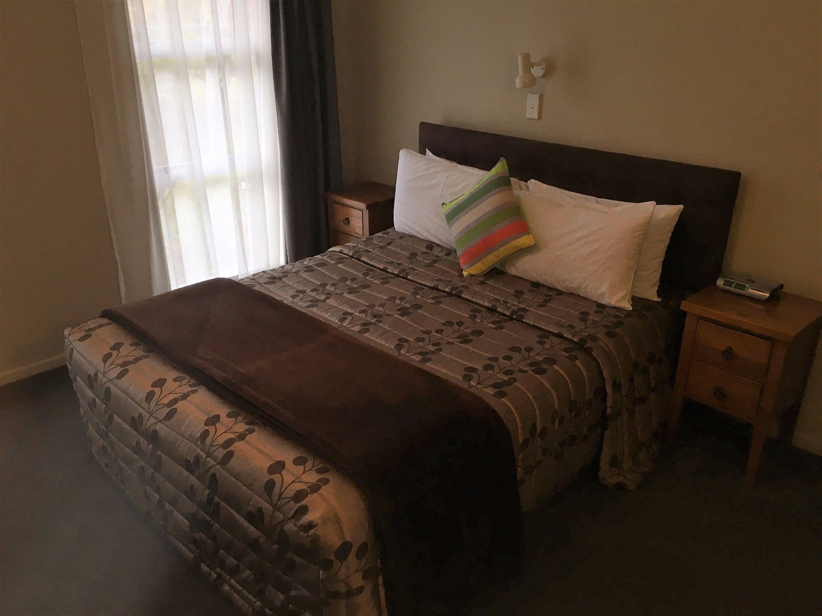 A neatly made bed with a patterned bedspread and pillows, a bedside table with a lamp, and a window with curtains.