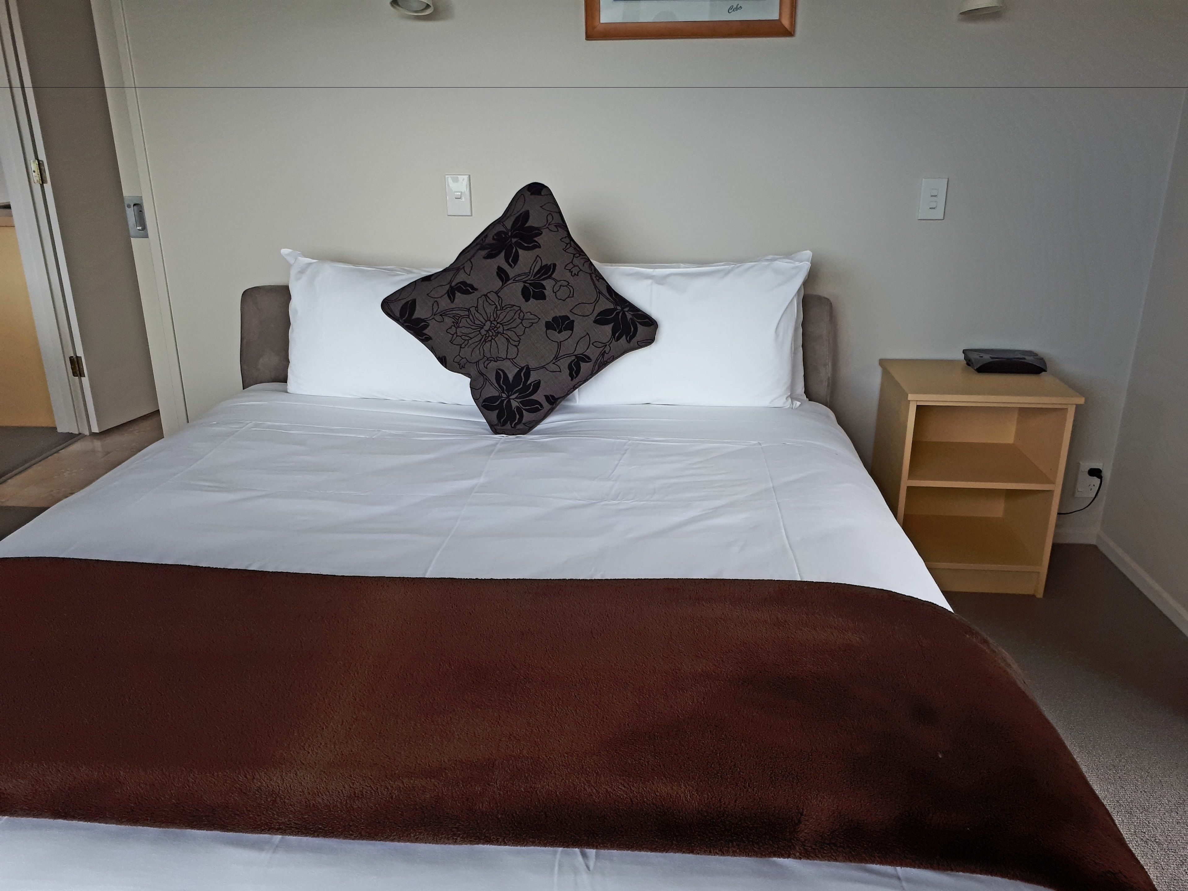 A neatly made bed with white linens and a brown throw blanket, a wooden nightstand with a lamp, and a framed picture on the wall.
