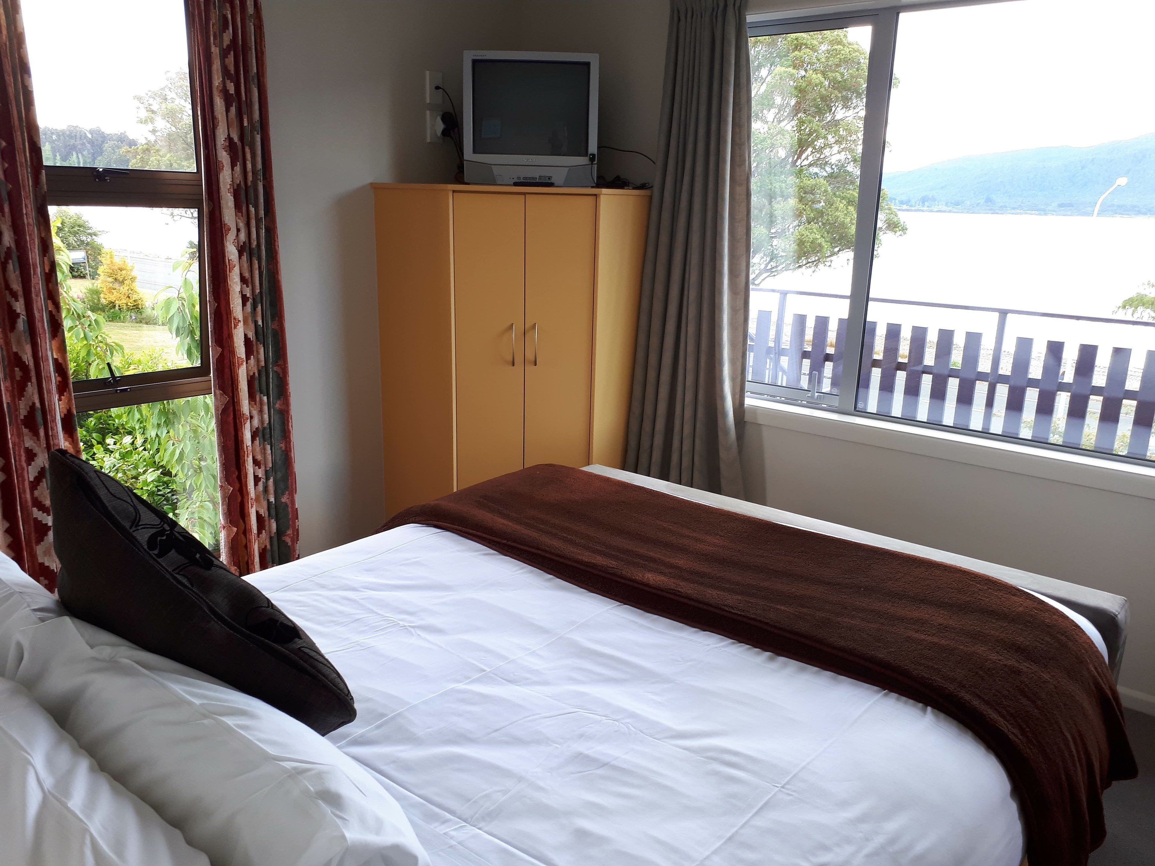 A hotel room with a bed, a television on a wooden cabinet, and a view of a lake through a window.