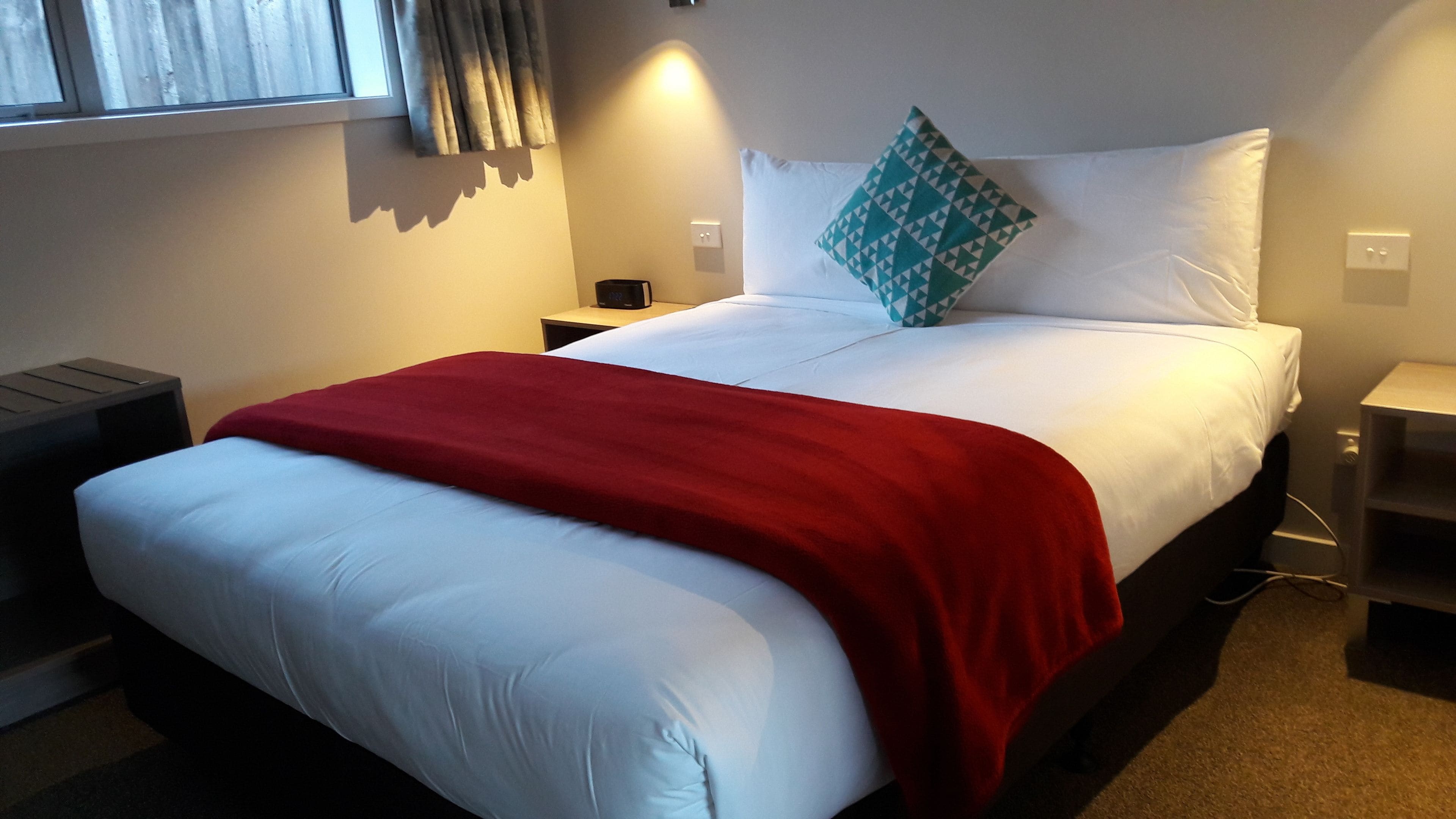 Quarto de hotel com cama, mesa de cabeceira, luminária e janela com cortinas.