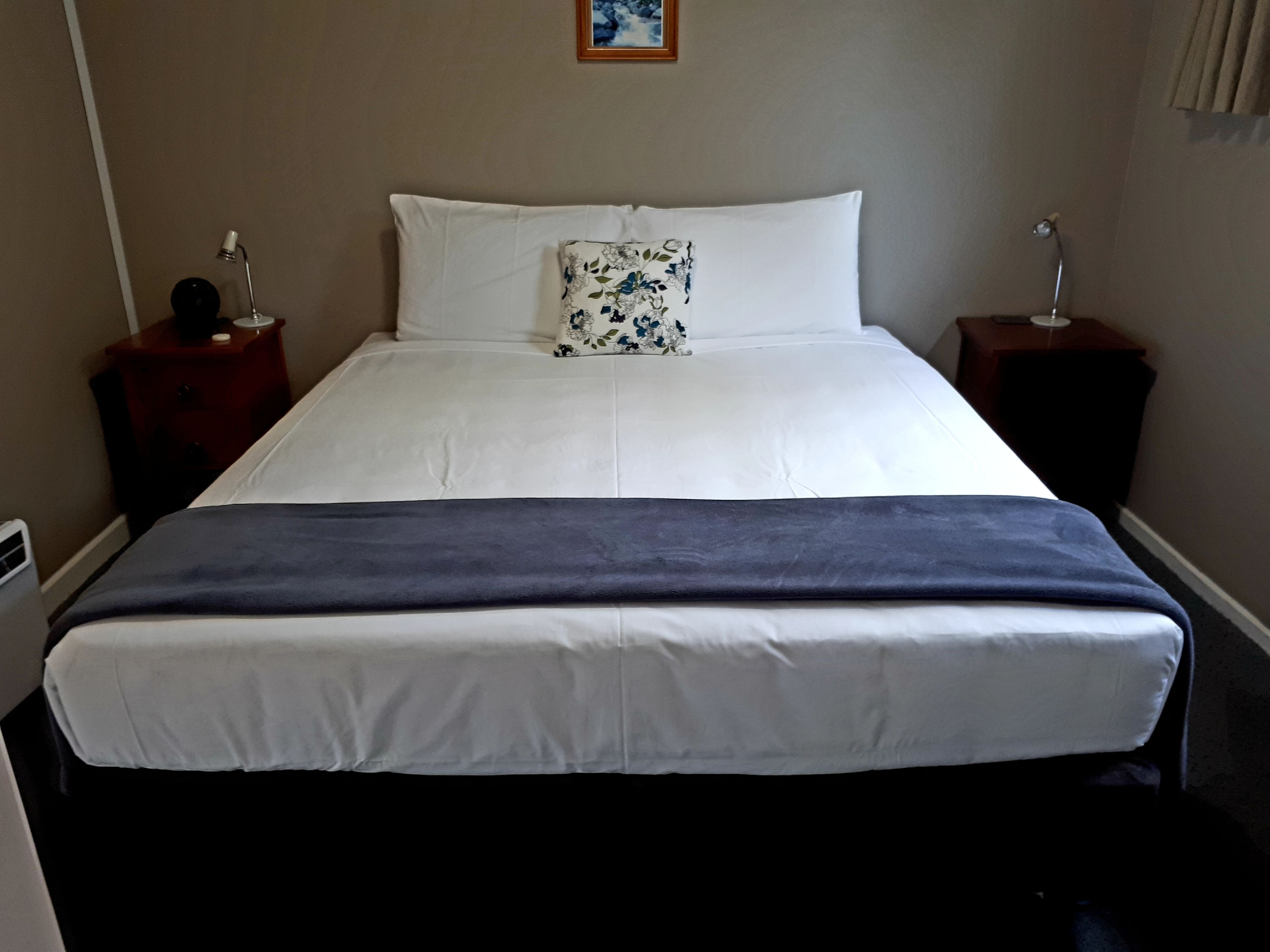 Una cama bien tendida con sábanas blancas y una manta azul, dos mesitas de noche con lámparas, y un cojín con estampado floral.