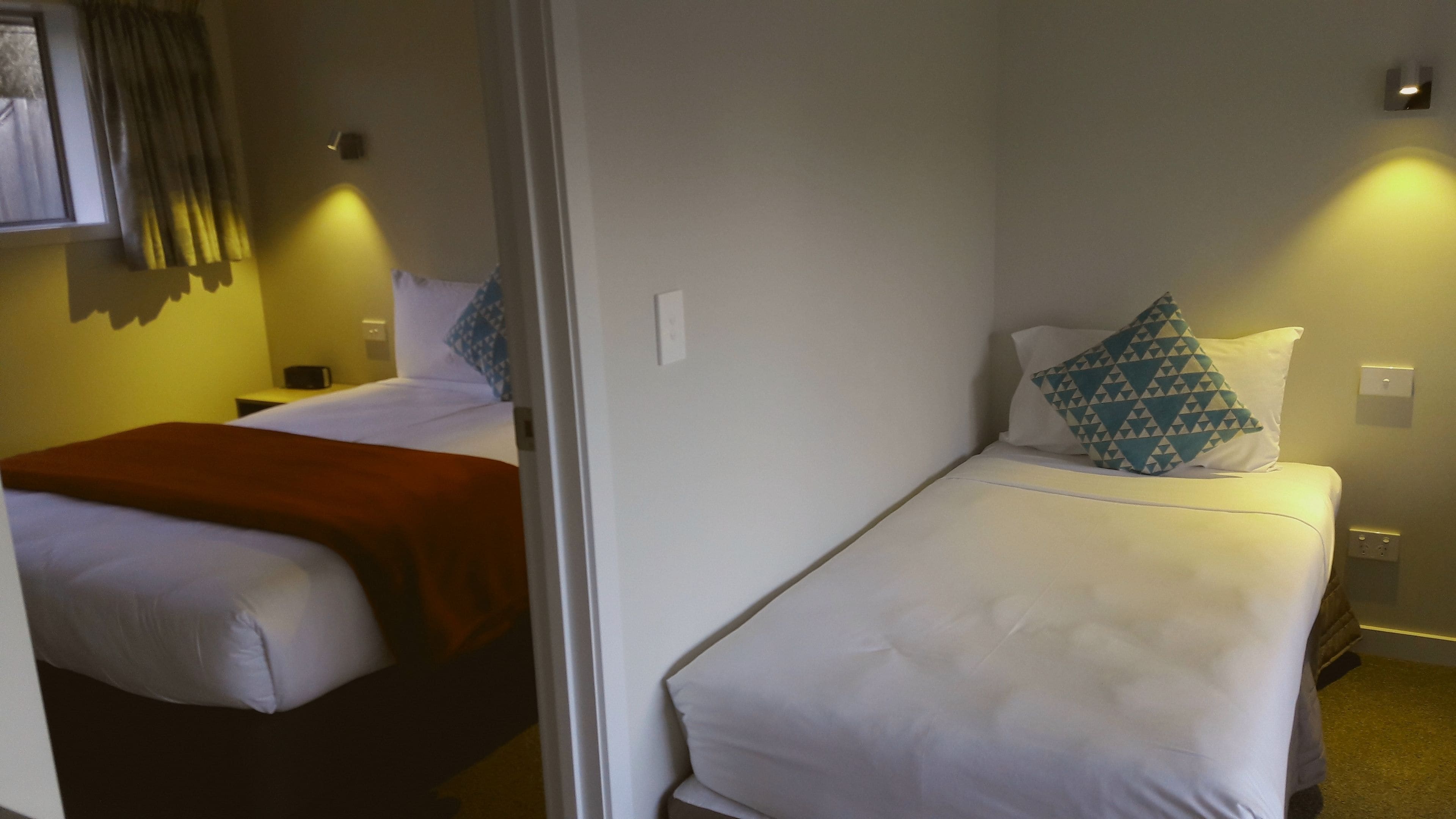 A hotel room with two beds, each with white bedding and a patterned pillow. There is a brown throw blanket on one bed. The room has a simple, functional design with minimal decor.