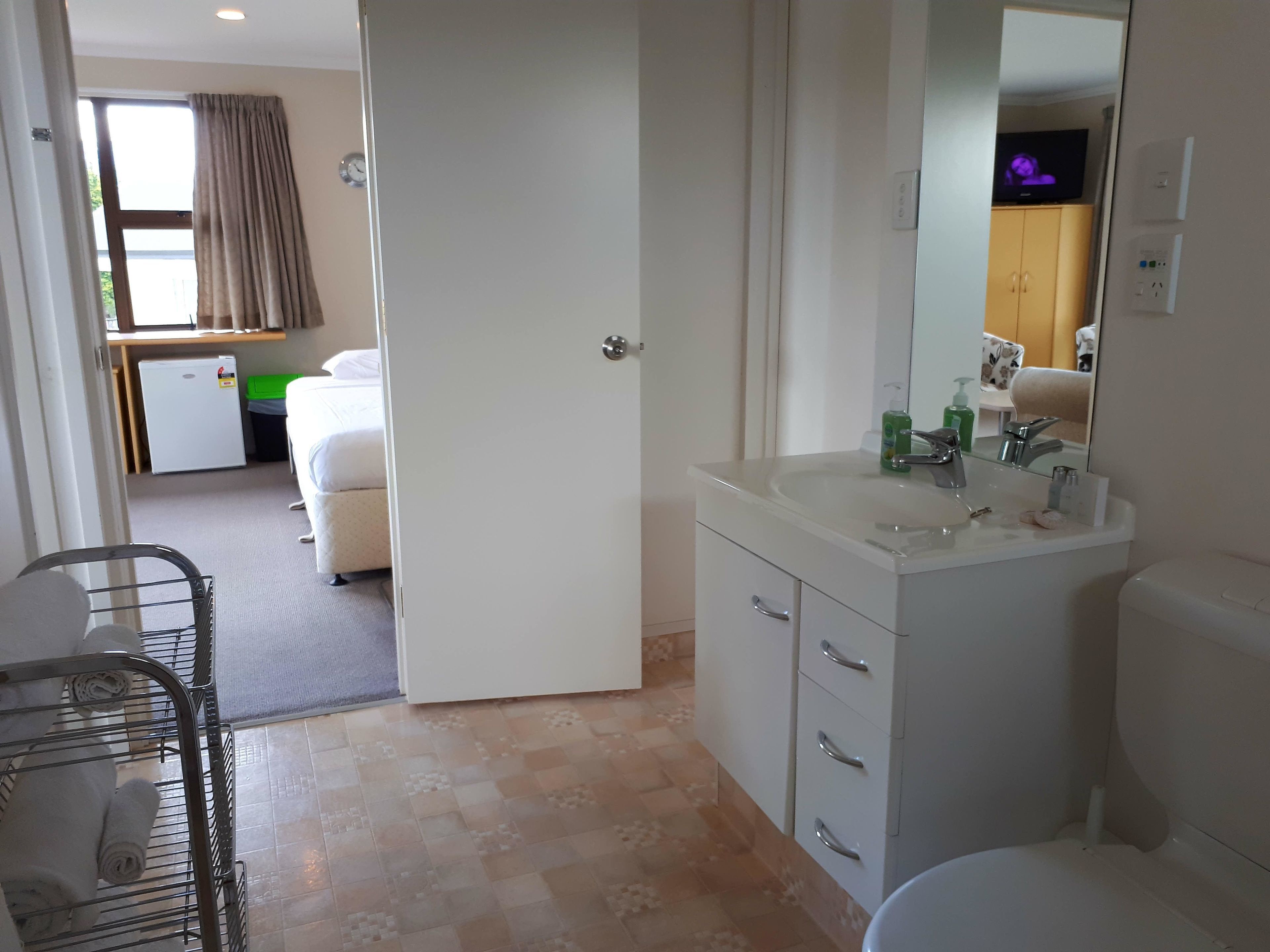 Une salle de bain d’hôtel avec un lavabo blanc, un miroir et des toilettes. Il y a une petite fenêtre avec des rideaux et une horloge au mur.