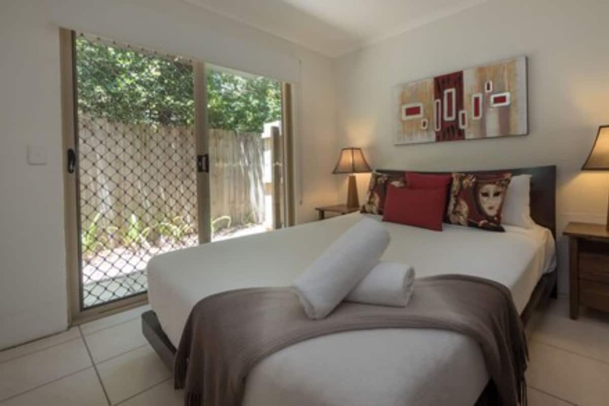 A hotel room with a bed, two bedside lamps, a large window with a view of greenery, and a painting on the wall.