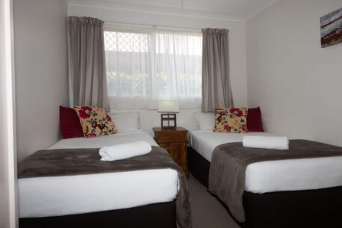 A hotel room with two single beds, a small wooden nightstand, a lamp, and a window with curtains.