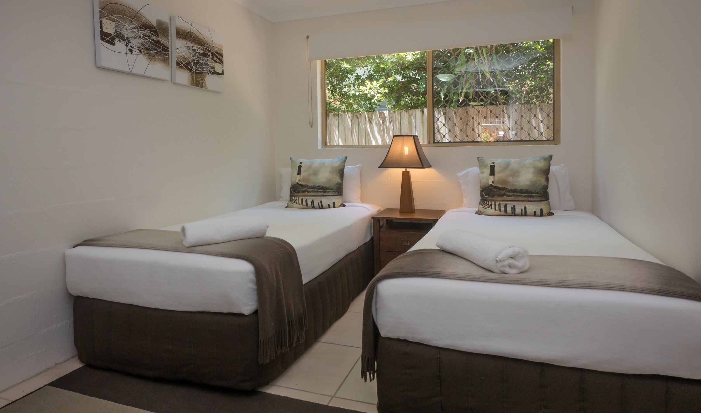 A hotel room with two single beds, a nightstand with a lamp, and artwork on the wall.