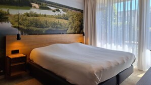 A bedroom with a large bed, a nightstand, a wall-mounted lamp, and a view of a lake through sheer curtains.